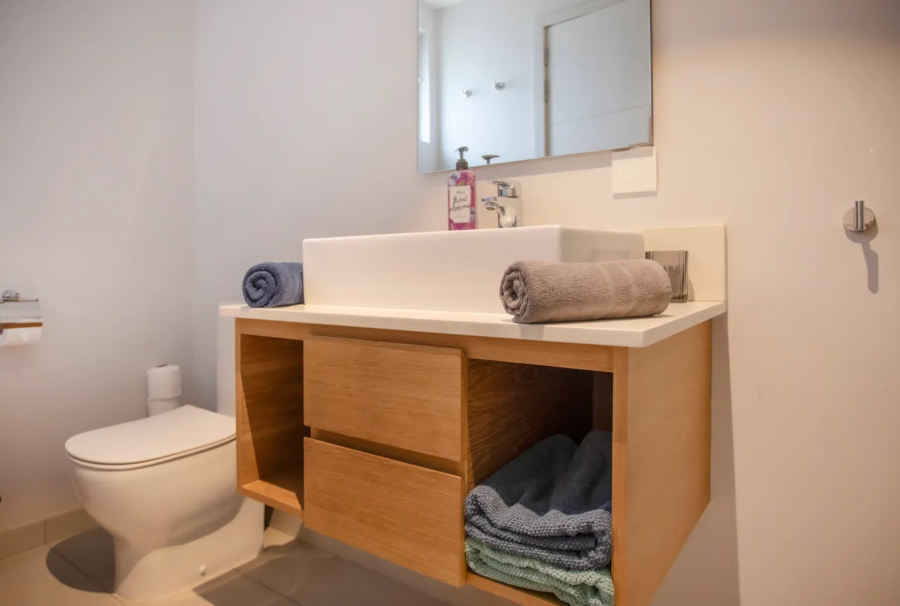 Salle de bain équipée d'un meuble en bois avec espace de rangement.  
Éclairage fonctionnel au-dessus du lavabo pour plus de confort.  
Douche moderne avec panneaux en verre et carrelage épuré.  
Linge de bain organisé sur un espace de rangement intégré.  
Sanitaire disposé de manière pratique pour un usage quotidien.  
Miroir encastré ajoutant une touche
