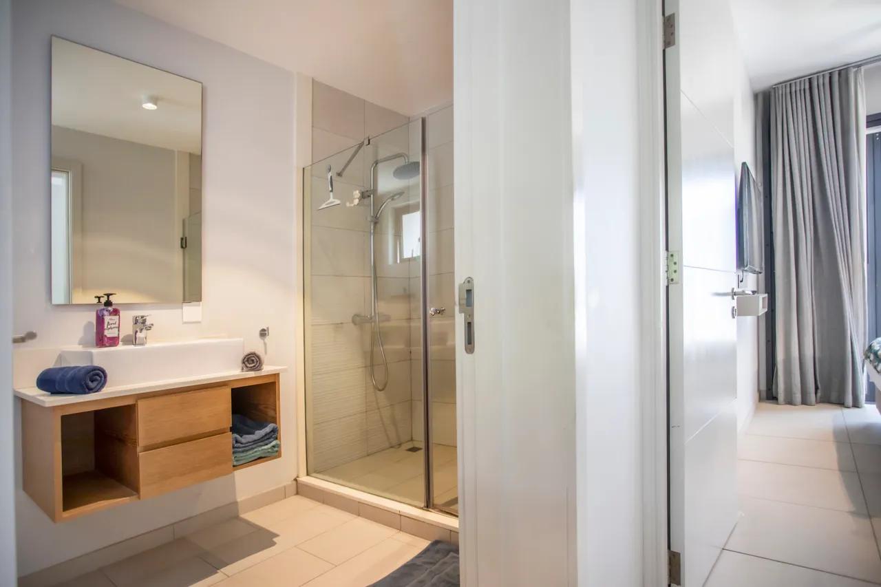 Salle de bain avec un meuble sous vasque en bois et miroir intégré.  
Douche spacieuse avec panneaux en verre et carrelage neutre.  
Éclairage au-dessus du miroir pour une utilisation pratique.  
Linge de bain soigneusement disposé sur un espace de rangement.  
Équipement moderne pour un confort quotidien dans la salle de bain.  
Fenêtre discrète offrant une