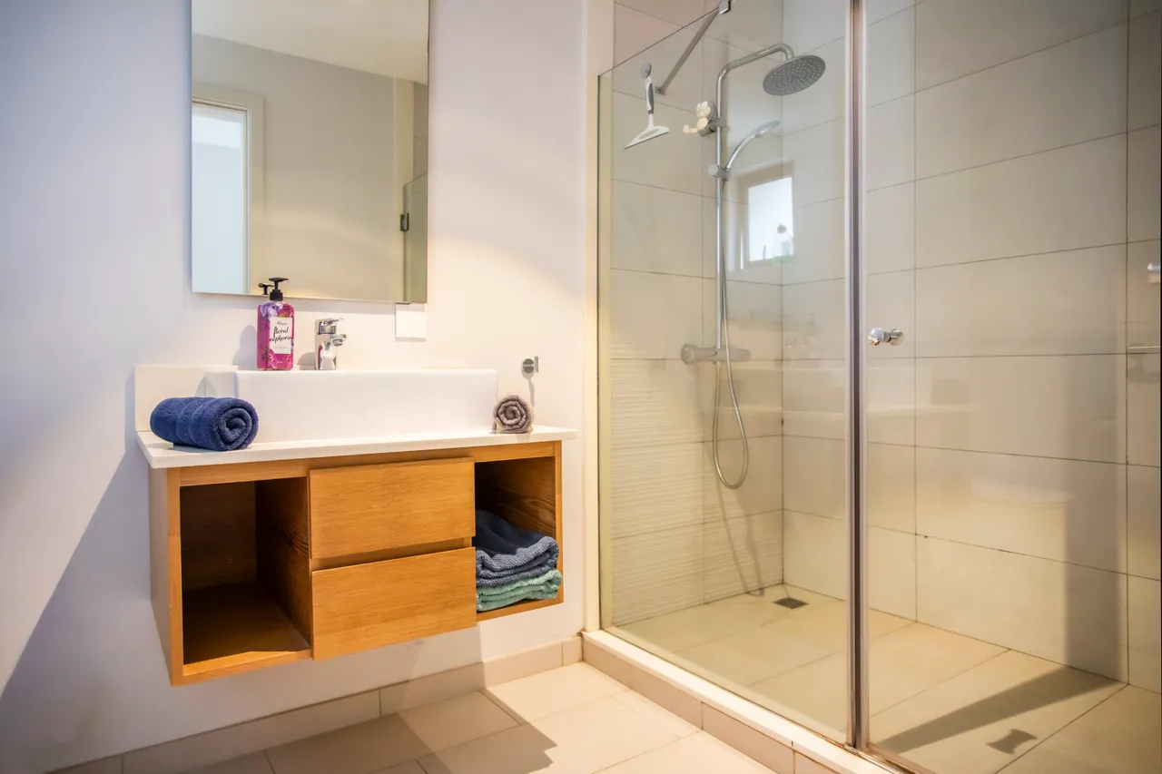 Douche moderne avec panneaux en verre et carrelage sobre.  
Éclairage fonctionnel placé au-dessus du miroir pour plus de commodité.  
Meuble sous vasque en bois offrant un espace de rangement pratique.  
Linge de bain organisé sur un espace de rangement intégré.  
Sanitaire disposé de manière pratique pour un usage quotidien.  
Miroir encastré ajoutant une