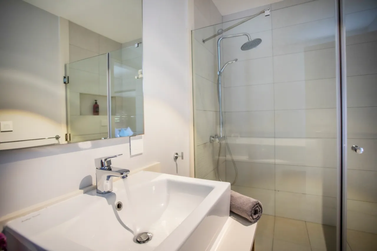 Salle de bain avec douche à l'italienne et parois en verre transparent.  
Meuble sous vasque en bois offrant un espace de rangement pratique.  
Éclairage fonctionnel placé au-dessus du miroir pour un usage quotidien.  
Linge de bain soigneusement disposé sur le meuble pour plus de commodité.  
Sanitaires bien agencés pour un accès fonctionnel.  
M