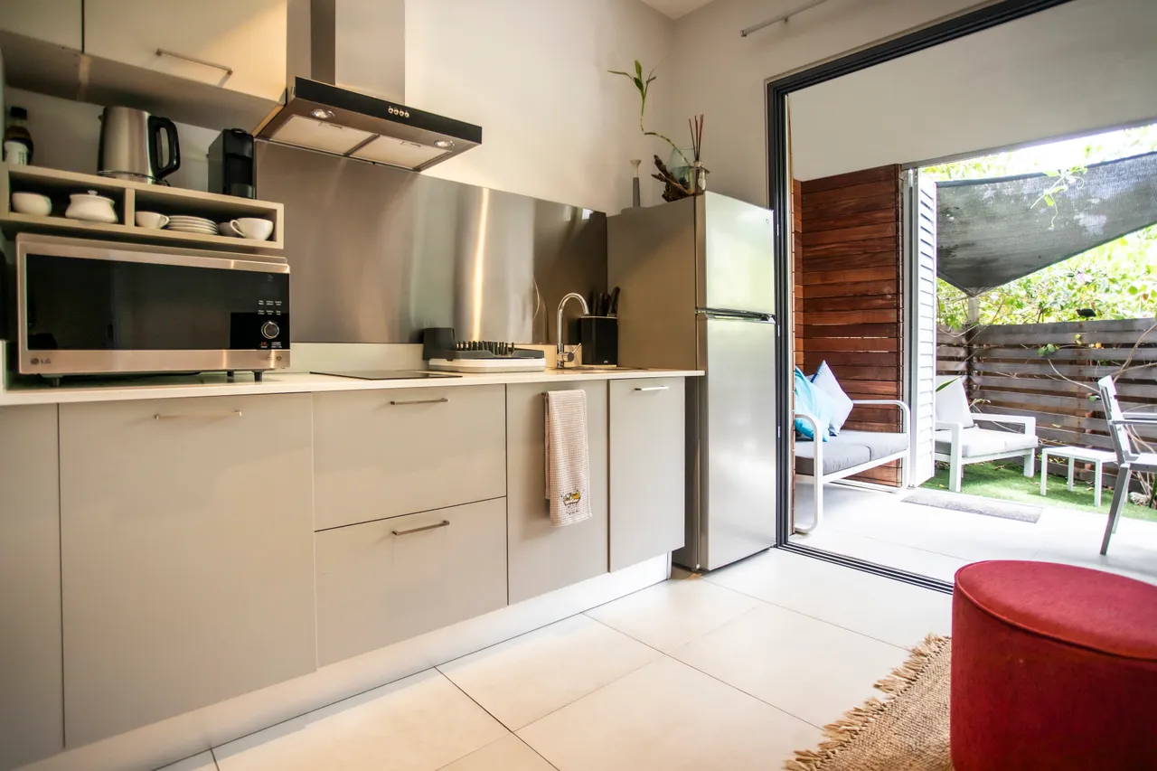Couleurs neutres et agencement moderne dans la cuisine fonctionnelle.  
Accès direct à une terrasse avec mobilier d'extérieur.  
Équipements électroménagers intégrés pour une utilisation pratique.  
Coin repas lumineux grâce à l’ouverture sur l’extérieur.  
Espace de rangement optimisé avec des placards modernes.  
Détails en bois ajoutant une touche naturelle à l'ensemble.