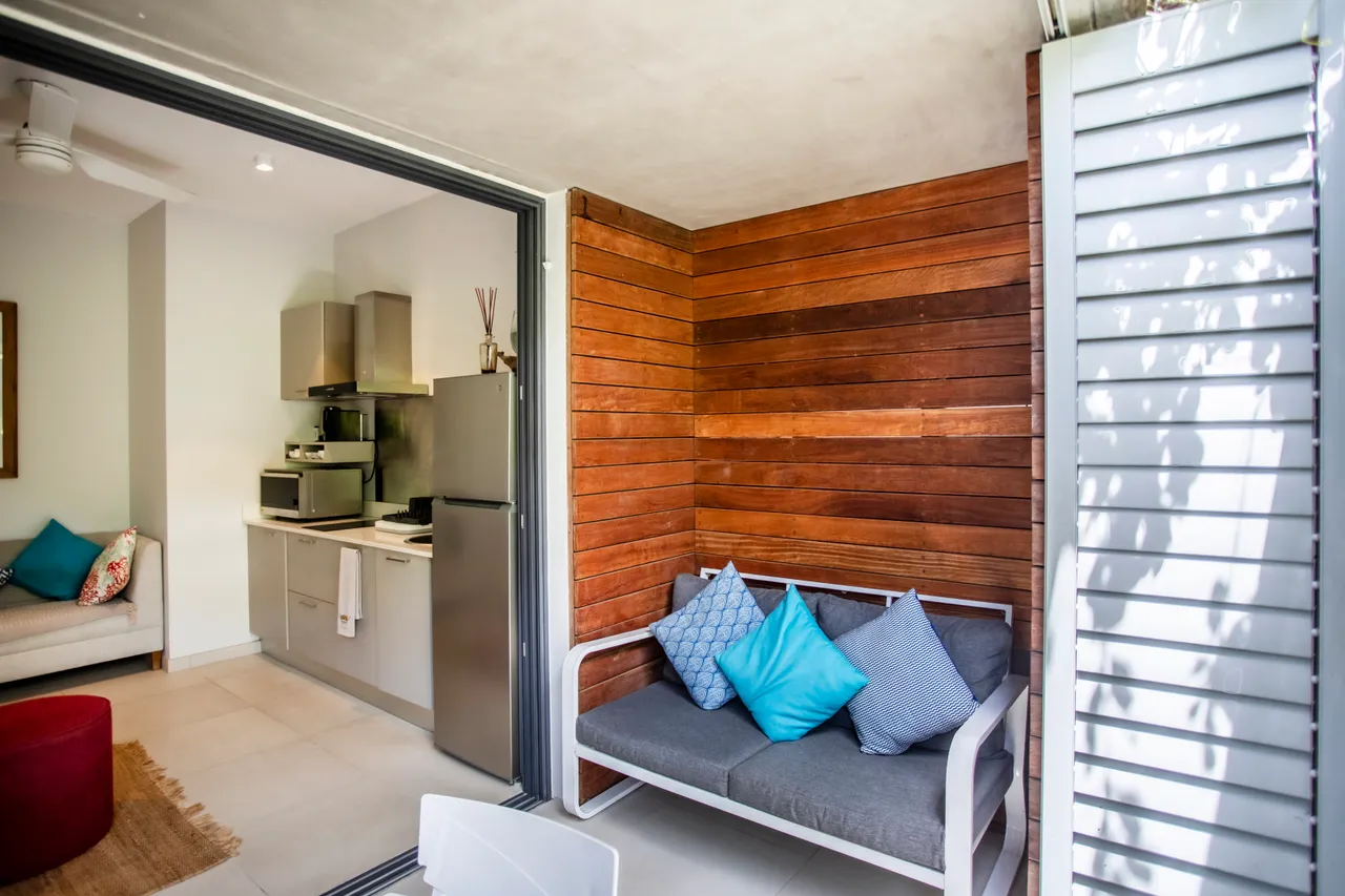 Espace de détente extérieur avec un canapé et des coussins colorés.  
Cuisine moderne avec réfrigérateur encastré et plan de travail fonctionnel.  
Ouverture vers l'extérieur offrant une vue sur l'environnement.  
Mur en bois apportant une touche chaleureuse à l'espace.  
Coin repas intégré avec un éclairage naturel agréable.