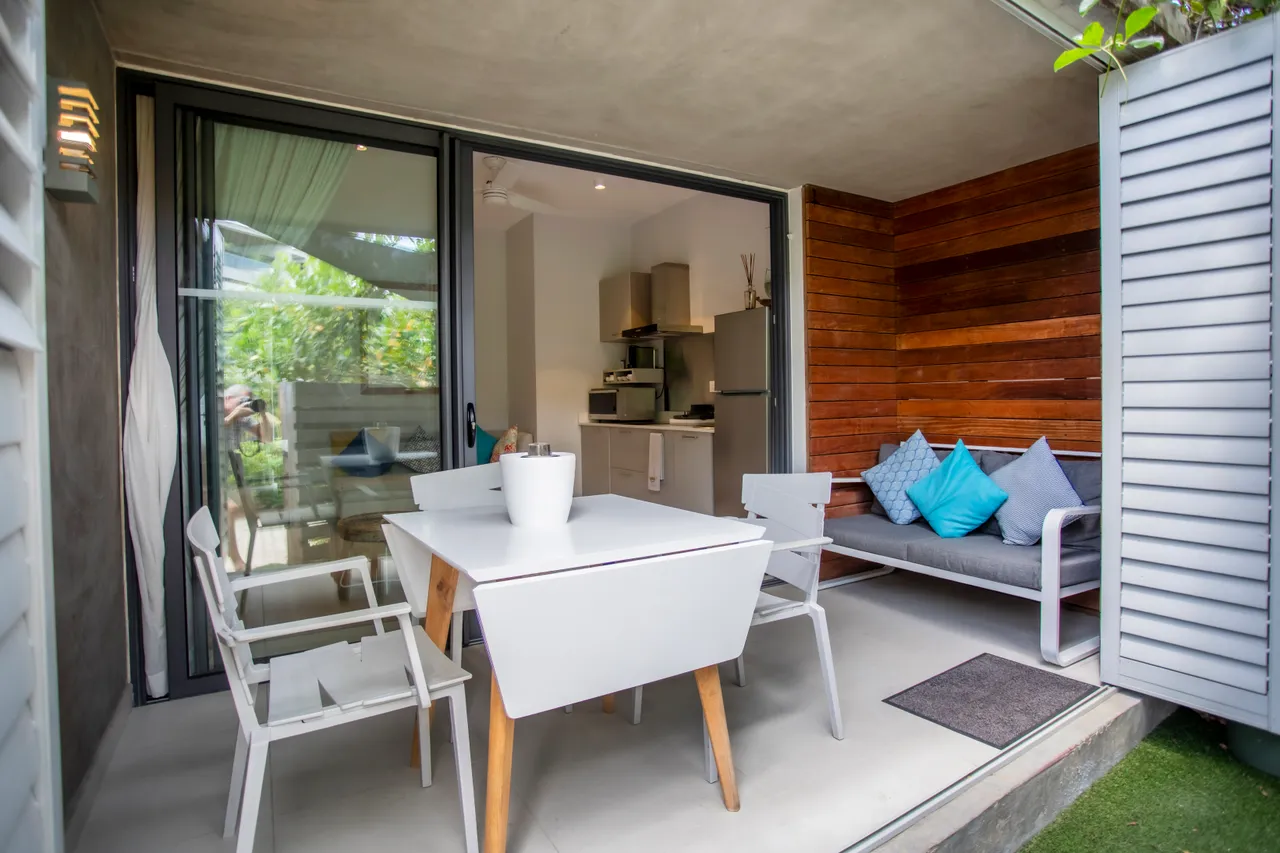 Espace extérieur avec table et chaises pour repas en plein air.  
Coin salon abrité avec canapé et coussins pour se détendre.  
Accès direct à la kitchenette pour plus de commodité.  
Terrasse avec sol en béton lisse, facile d'entretien.  
Végétation environnante offrant une atmosphère tranquille et intime.