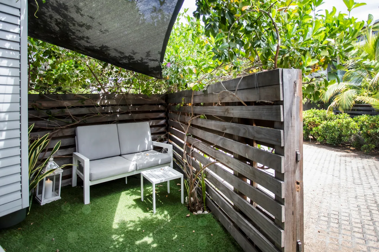 Espace extérieur privé avec canapé confortable et petite table.  
Végétation dense offrant une certaine intimité pour se détendre.  
Sol en gazon synthétique facilitant l'entretien de la zone.  
Clôture en bois créant une séparation discrète tout en restant ouverte.  
Coin ombragé idéal pour profiter de l'extérieur en toute tranquillité.