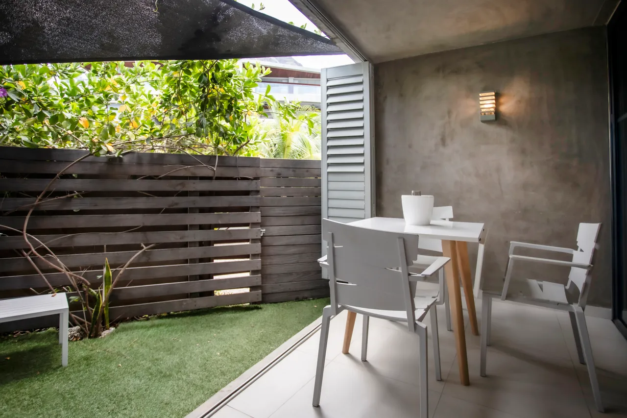 Espace extérieur avec table et chaises blanches pour repas à l'air libre.  
Végétation environnante ajoutant une touche de verdure à l'espace.  
Panneau en bois créant une séparation discrète et esthétique.  
Sol en gazon synthétique offrant un entretien minimal.  
Éclairage mural fonctionnel pour profiter de l'extérieur en soirée.
