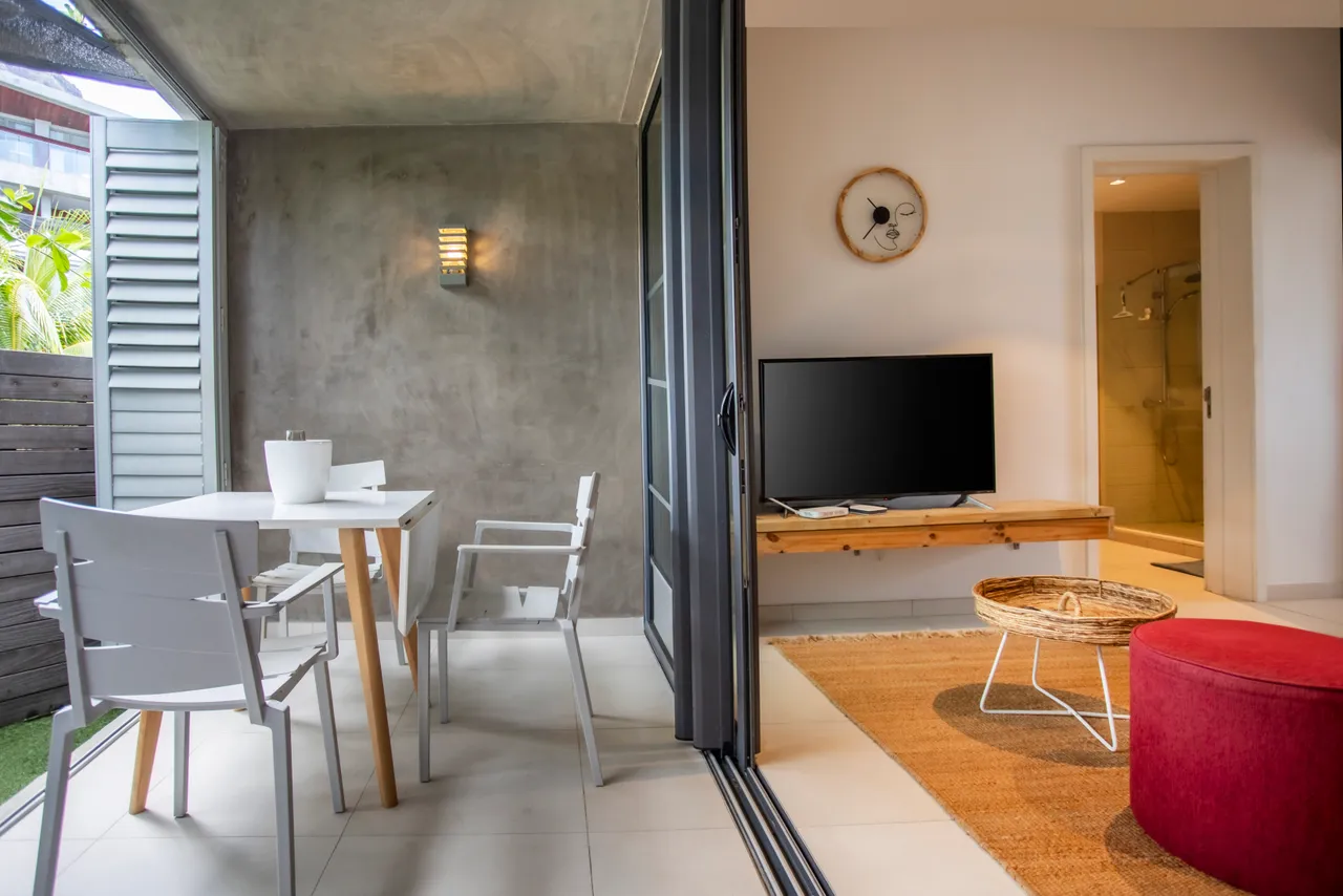 Espace de vie lumineux avec télévision et meubles modernes.  
Coin repas avec table et chaises, offrant une vue agréable.  
Sol carrelé facilitant l'entretien et ajoutant une touche contemporaine.  
Ouverture sur un espace extérieur verdoyant pour plus de lumière.  
Détails décoratifs sur les murs ajoutant une note artistique à l'ensemble.