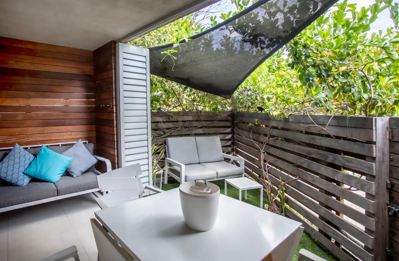 Espace extérieur avec mobilier moderne et coussins colorés pour se détendre.  
Végétation environnante créant une ambiance intime et apaisante.  
Sol en gazon synthétique facilitant l'entretien de la zone.  
Pare-soleil offrant un abri tout en laissant passer la lumière.  
Table blanche et chaises assorties pour des repas en plein air.