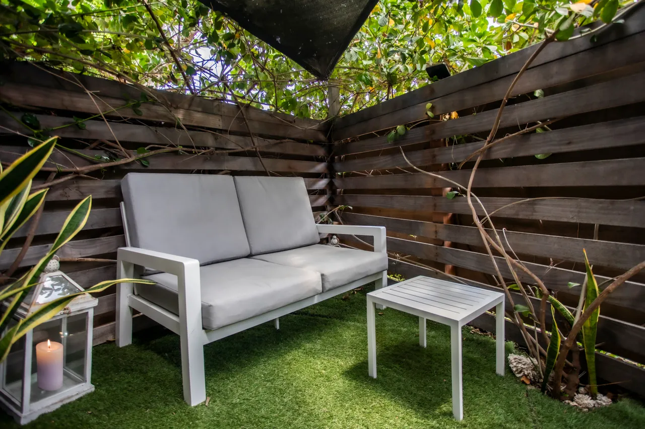 Espace détente extérieur avec canapé et table basse sur gazon synthétique.  
Végétation dense créant une ambiance intime et agréable.  
Clôture en bois ajoutant une séparation tout en restant légère.  
Coin ombragé pour profiter du calme en plein air.