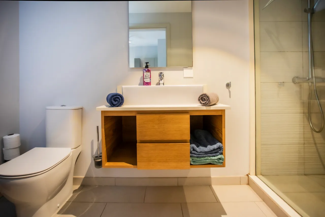 Salle de bain équipée d'un meuble sous vasque en bois et d'une douche.  
Étagères ouvertes pour un rangement pratique des serviettes et accessoires.  
Miroir mural ajoutant une touche de lumière à l'espace.  
Sol carrelé facilitant l'entretien tout en restant sobre et contemporain.  
Sanitaire moderne assurant confort et fonctionnalité dans la pièce.