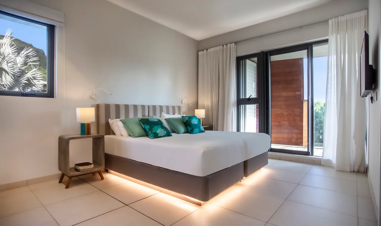 Tons neutres et murs clairs créent une atmosphère apaisante. Chambre équipée d'un lit king-size.  
Luminosité naturelle grâce aux grandes fenêtres offrant une vue partielle sur l'extérieur.  
Table de chevet moderne en bois avec éclairage intégré, ajoutant une touche fonctionnelle.  
Coussins décoratifs aux teintes turquoise, apportant une note de couleur