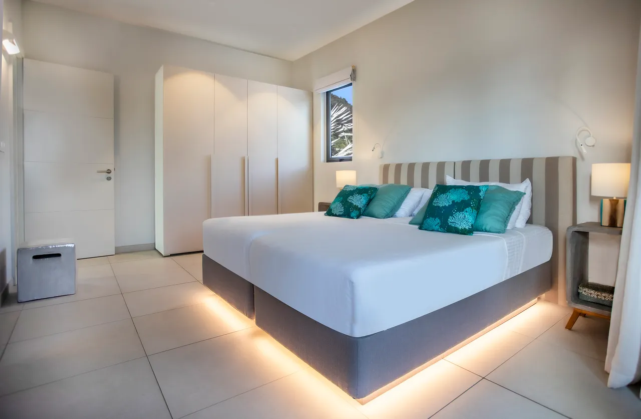 Chambre avec grandes fenêtres offrant une vue sur le jardin. Éclairage doux sous le lit. Mobilier moderne avec têtes de lit assorties. Rangement ample grâce à des placards intégrés.