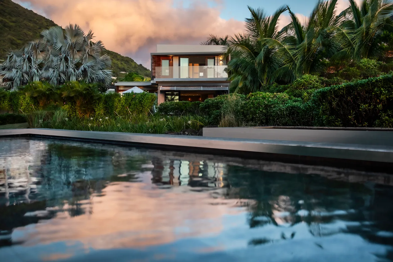 Terrasse en bois autour de la piscine, offrant un accès direct au jardin. Végétation tropicale luxuriante encadre l'espace extérieur. Réflexion du ciel coloré dans l'eau de la piscine.