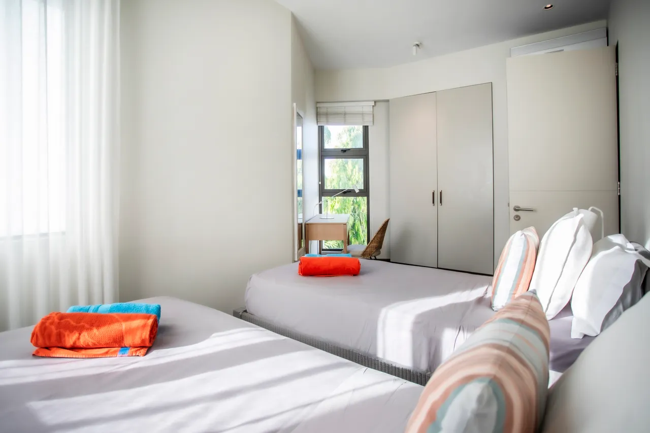 Chambre avec deux lits simples et rangement discret. Éclairage naturel et textile coloré.