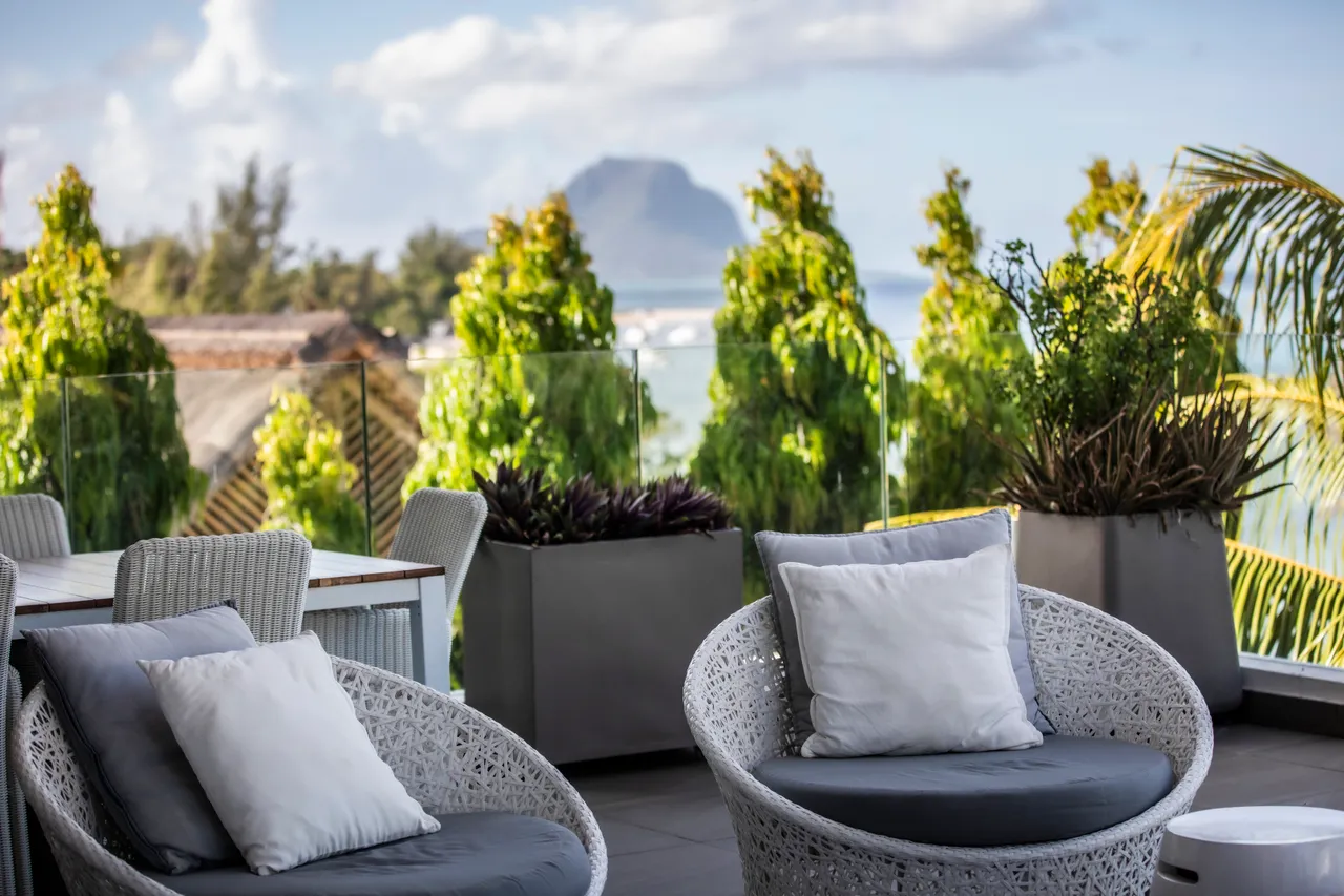 Terrasse avec fauteuils en rotin et vue sur la mer et les montagnes. Plantes vertes ajoutent une touche naturelle à l'espace.