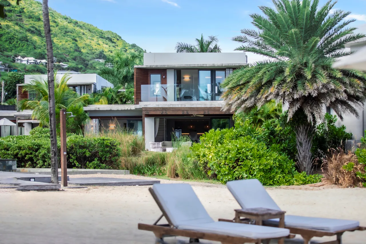 Villa en bord de mer avec accès direct à la plage.  

Terrasse surélevée offrant une vue sur le jardin et la mer.  

Végétation tropicale entourant la villa pour plus d'intimité.  

Mobilier extérieur en bois avec chaises longues près du sable.  

Architecture moderne intégrée dans un cadre naturel verdoyant.