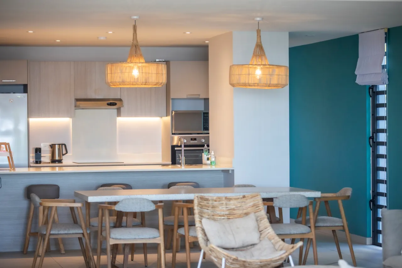Espace repas avec table en bois et chaises confortables.  

Lumière naturelle grâce aux grandes fenêtres orientées vers l'extérieur.  

Éclairage suspendu en rotin ajoutant une touche moderne à l'espace.  

Disposition ouverte facilitant la circulation vers la cuisine.  

Sol carrelé clair renforçant l'aspect contemporain de la pièce.