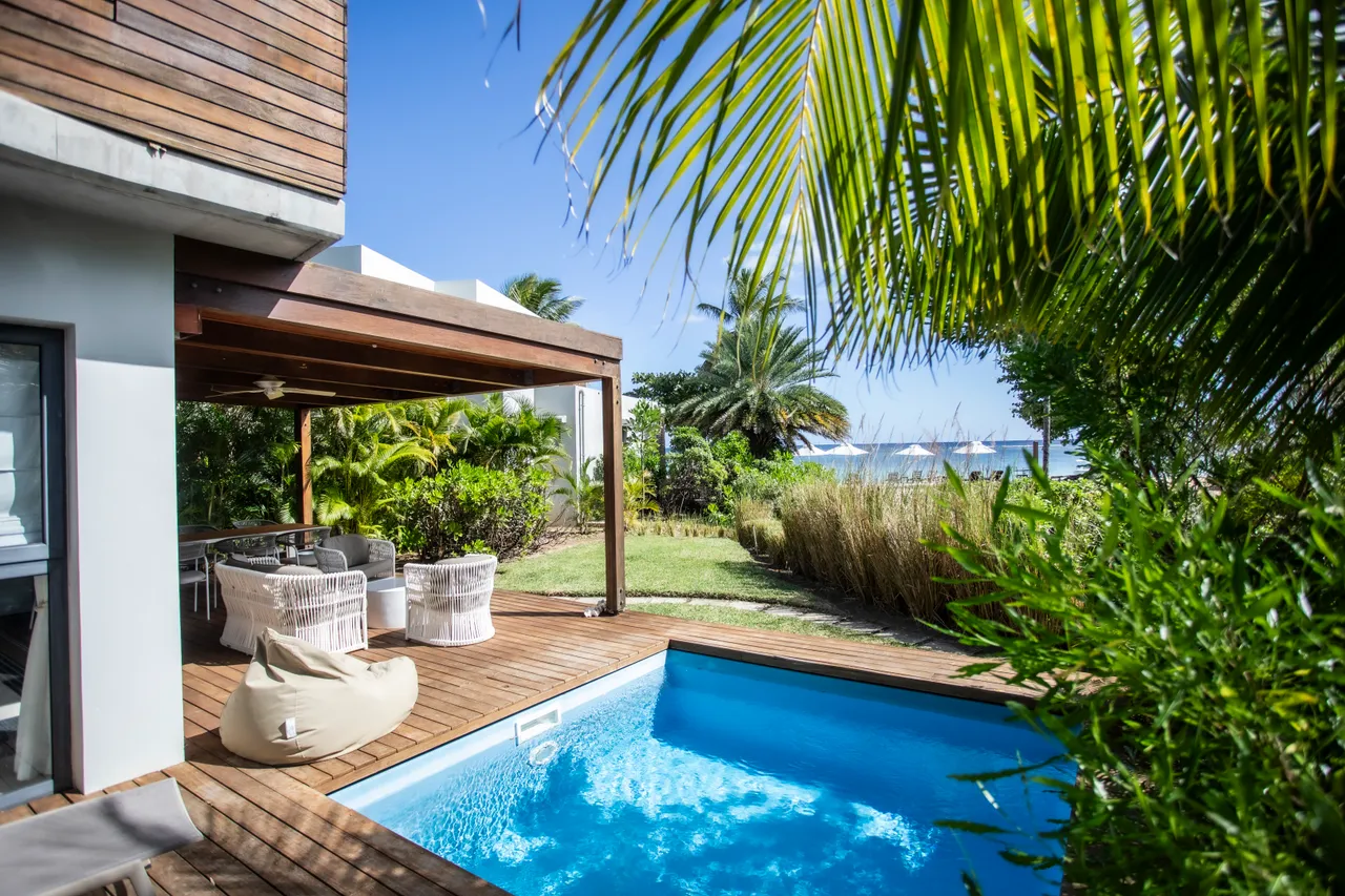 Terrasse en bois avec coin repas offrant une vue sur la piscine.  

Chaises longues en bois disposées autour de la piscine privée.  

Végétation tropicale entourant l'espace extérieur pour plus d'intimité.  

Accès direct à la plage depuis le jardin verdoyant.  

Espace détente avec mobilier en rotin et ambiance sereine.