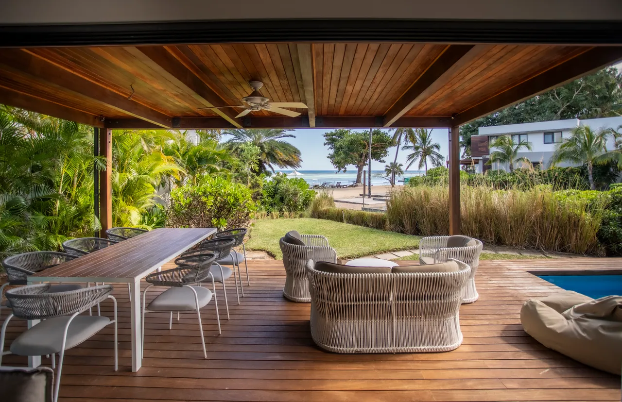 Espace extérieur couvert avec une vue sur la mer et le jardin.  

Mobilier en rotin pour un coin salon confortable et fonctionnel.  

Table en bois pour des repas en plein air.  

Végétation tropicale créant une ambiance sereine autour de l'espace.  

Accès direct à la piscine et à la plage depuis la terrasse.