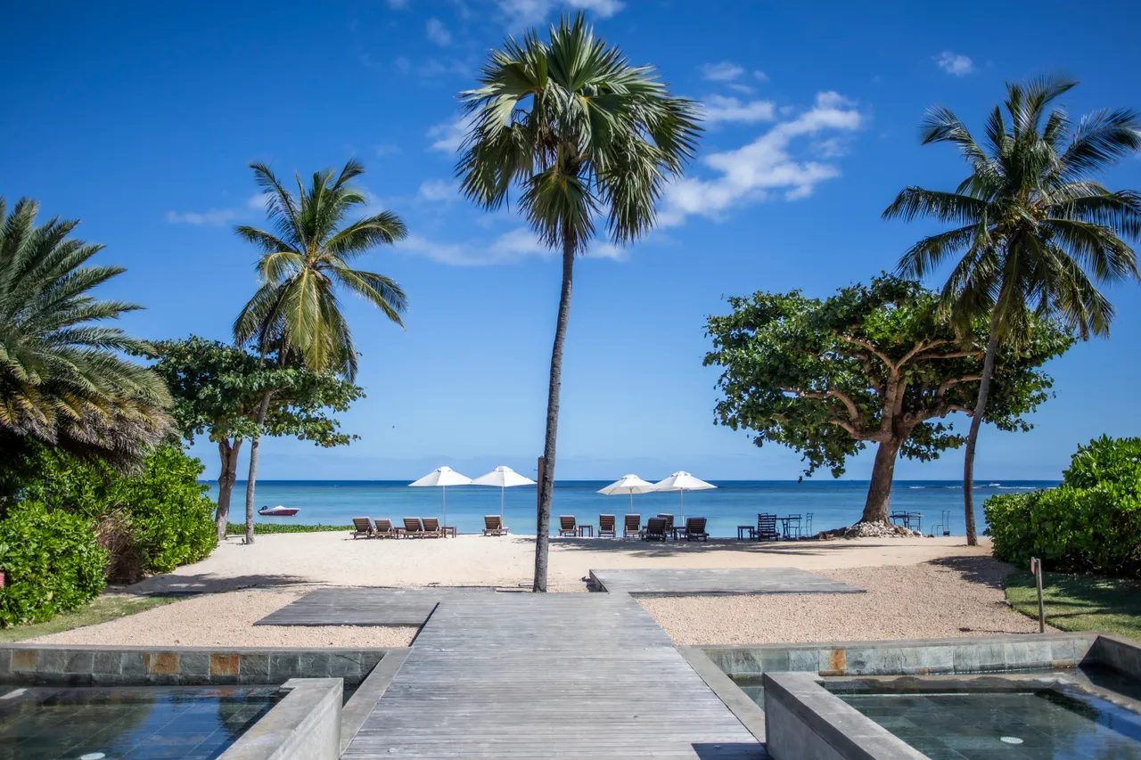 Accès direct à la plage depuis le jardin privatif.  

Palmiers et parasols blancs bordant l'espace de sable.  

Espace de baignade aménagé avec chaises longues sur le sable.  

Vue dégagée sur l'océan depuis la terrasse en bois.  

Végétation dense offrant de l'ombre près de la plage.