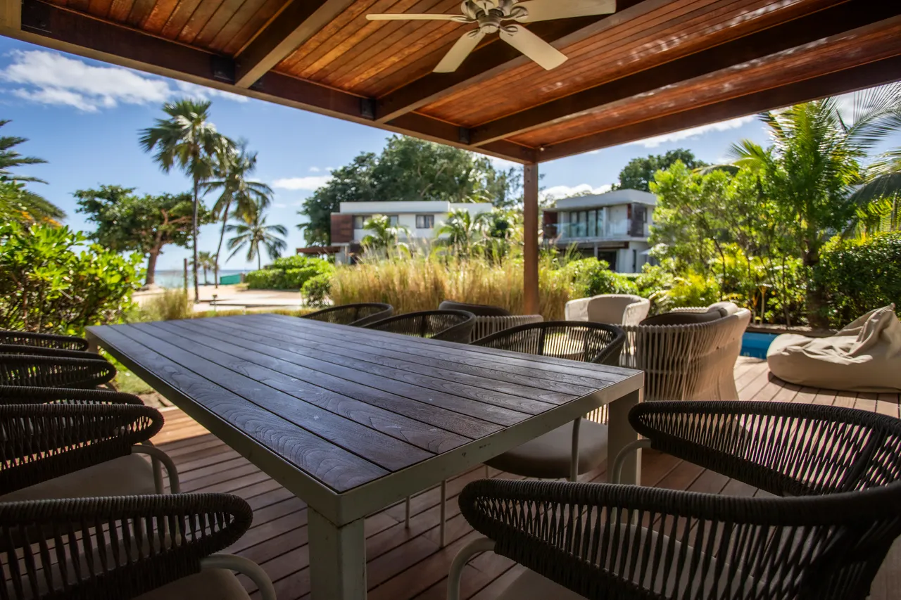 Espace repas abrité avec table en bois et chaises confortables.  

Vue sur le jardin verdoyant et la plage à proximité.  

Plafond en bois créant une atmosphère naturelle et accueillante.  

Éclairage naturel grâce aux larges ouvertures sur l'extérieur.  

Mobilier en rotin ajoutant une touche décontractée à l'espace.