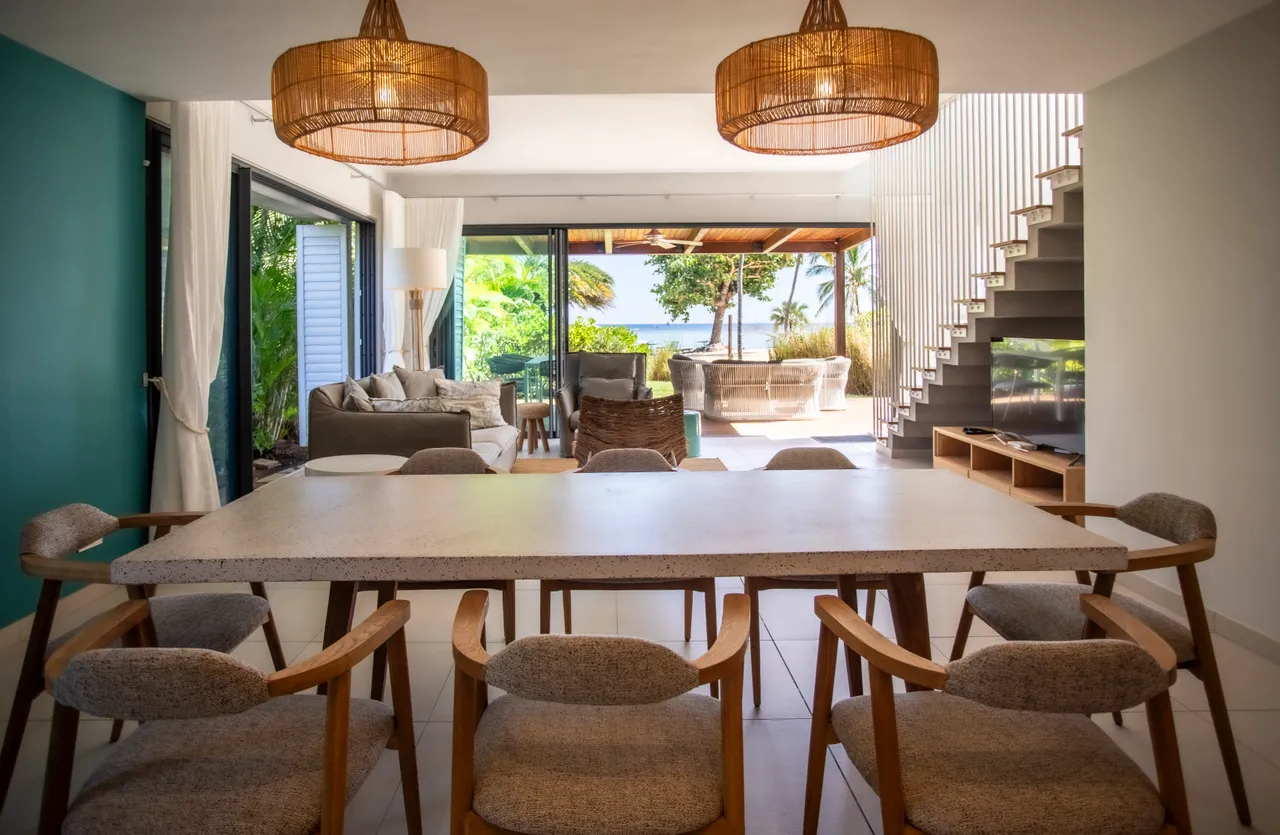 Espace repas avec grandes fenêtres offrant une vue sur le jardin.  

Mobilier en bois clair et éclairage suspendu en rotin naturel.  

Disposition ouverte reliant la salle à la terrasse extérieure.  

Ambiance accueillante avec des couleurs apaisantes et contemporaines.  

Sol carrelé clair ajoutant une touche moderne à l'espace.