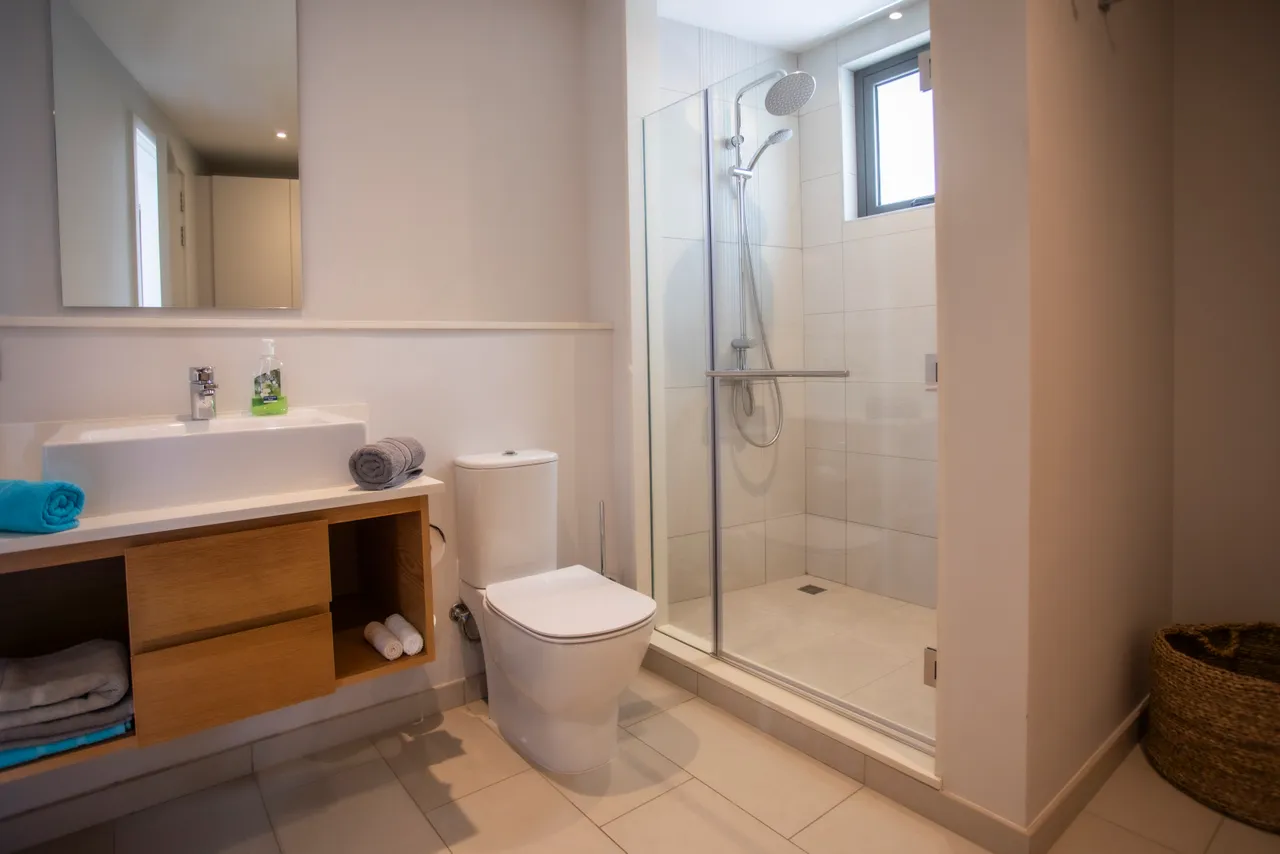 Salle de bain lumineuse avec douche vitrée et mobilier en bois clair.  

Rangement pratique sous le lavabo pour serviettes et accessoires.  

Surface carrelée beige offrant une ambiance neutre et moderne.  

Éclairage naturel grâce à la fenêtre intégrée au design.  

Accessoires de bain soigneusement disposés pour un espace organisé.