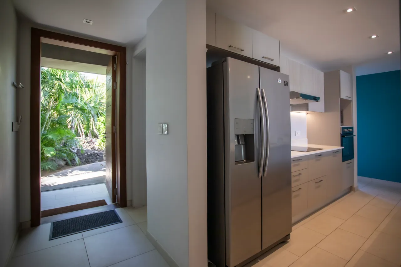 Mur bleu accentuant la cuisine ouverte et moderne. 

Réfrigérateur en acier inoxydable intégré pour un usage pratique.  

Plans de travail clairs, facilitant la préparation des repas.  

Lumière naturelle inondant l'espace grâce à l'accès extérieur.  

Rangements fonctionnels en dessous du plan de travail.