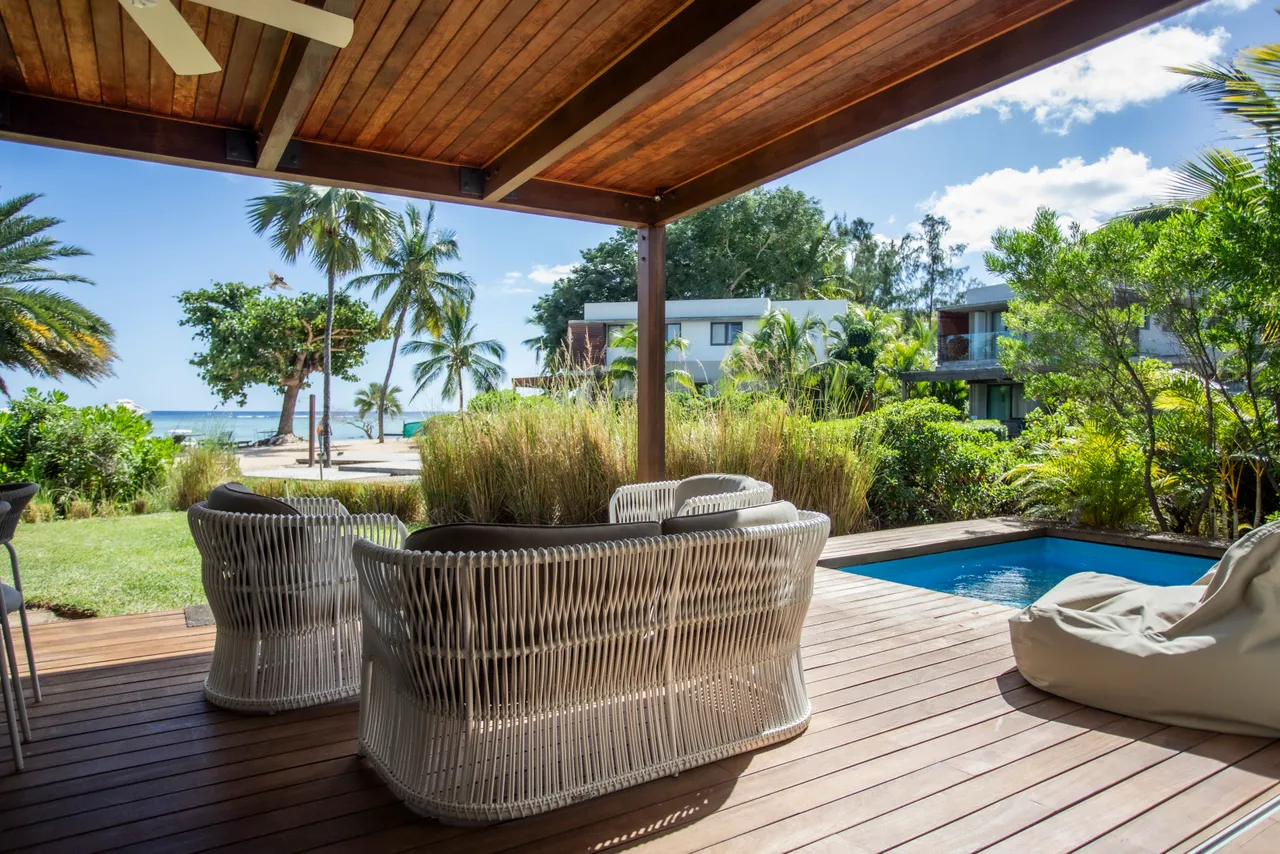 Terrasse couverte avec mobilier en rotin, offrant une vue sur la mer.  

Espace dehors agrémenté de verdure et d'une petite piscine.  

Lumière naturelle filtrant à travers le toit en bois.  

Coin salon extérieur propice à la détente près de la plage.  

Végétation tropicale créant une ambiance calme et naturelle.