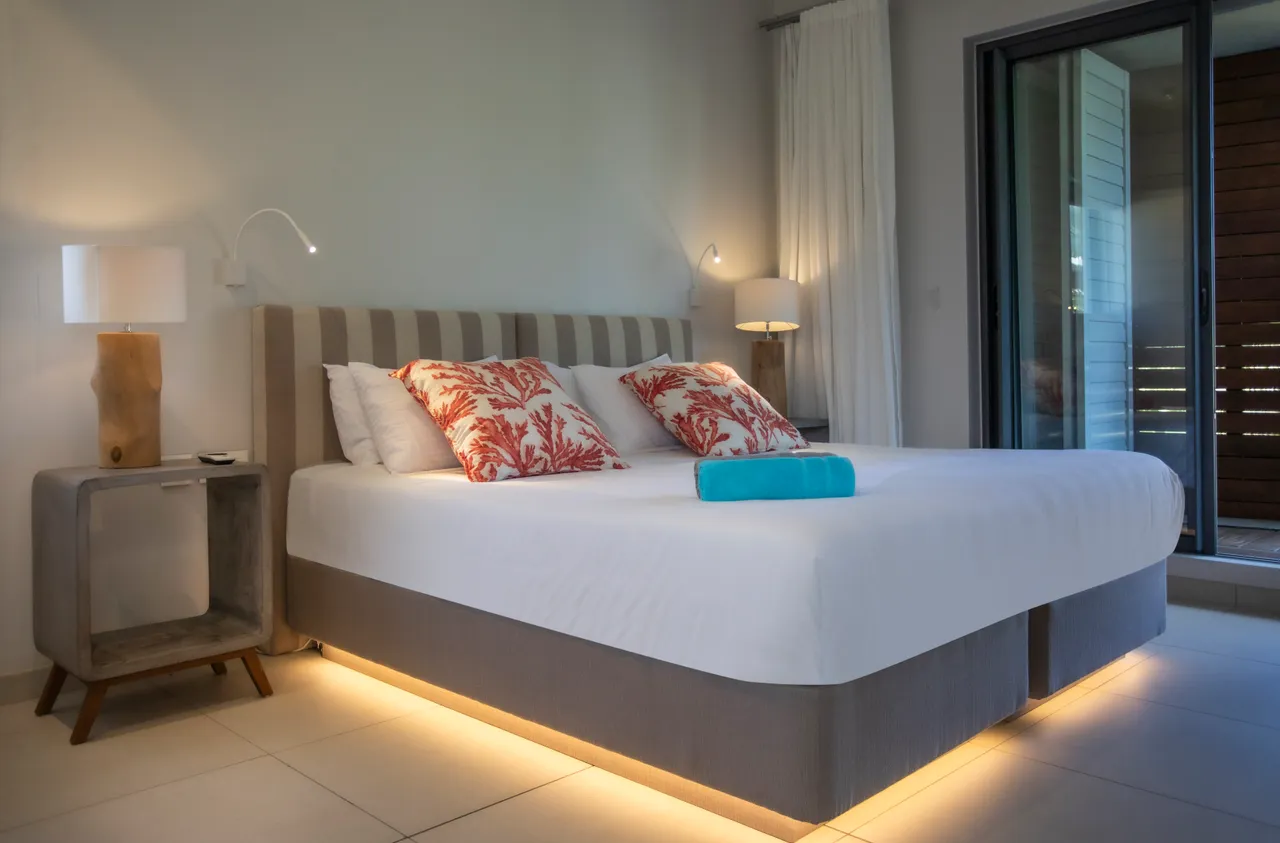Chambre avec lit double et accès à une terrasse privée.  

Linge de lit clair et coussins colorés ajoutant une touche de couleur.  

Éclairage indirect soulignant le design contemporain et apaisant.  

Mobilier minimaliste et fonctionnel pour un espace reposant.  

Fenêtre offrant une vue sur l'extérieur et favorisant la lumière naturelle.