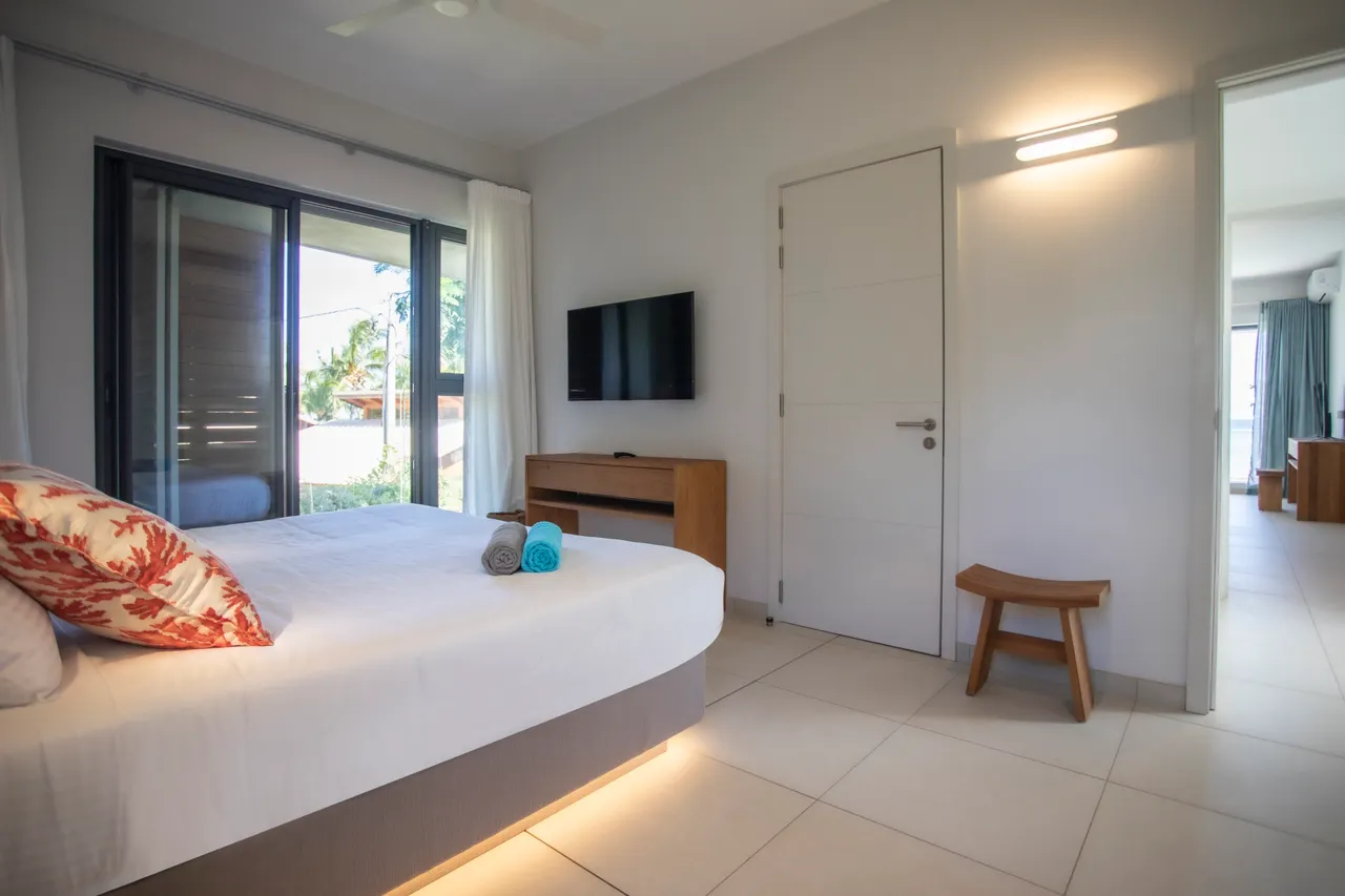 Chambre avec lit double et télévision murale pour un confort moderne.  

Éclairage doux créant une atmosphère accueillante et reposante.  

Fenêtres offrant une vue sur l'extérieur et une bonne luminosité.  

Mobilier simple en bois ajoutant une touche naturelle à l'espace.  

Accès direct à une salle de bain attenante pour plus de commodité.
