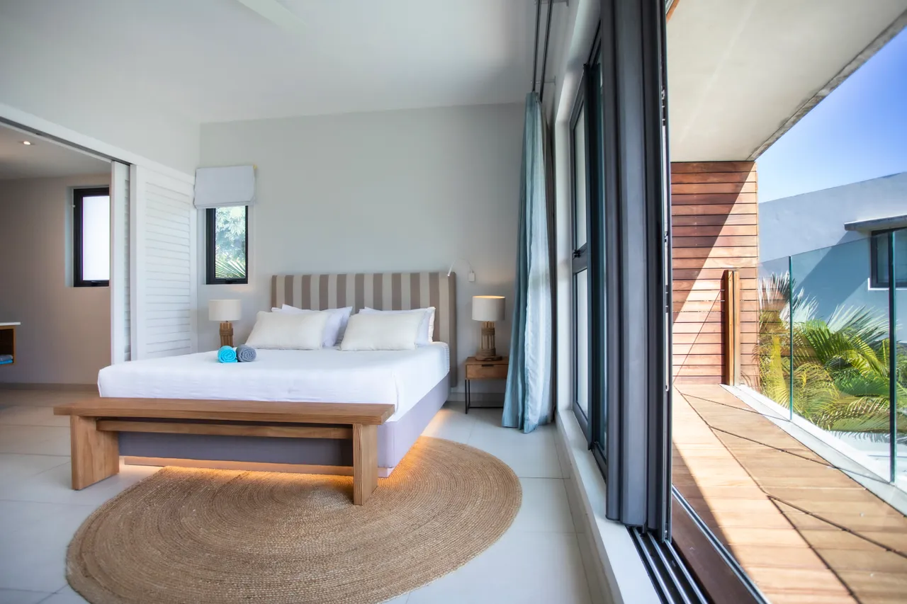 Chambre avec lit double et accès à une terrasse en bois.  

Fenêtres offrant une vue sur le jardin tropical et la piscine.  

Mobilier contemporain avec éclairage indirect créant une ambiance douce.  

Tapis en jute ajoutant une texture naturelle à l'espace.  

Rangement intégré pour un environnement dégagé et fonctionnel.