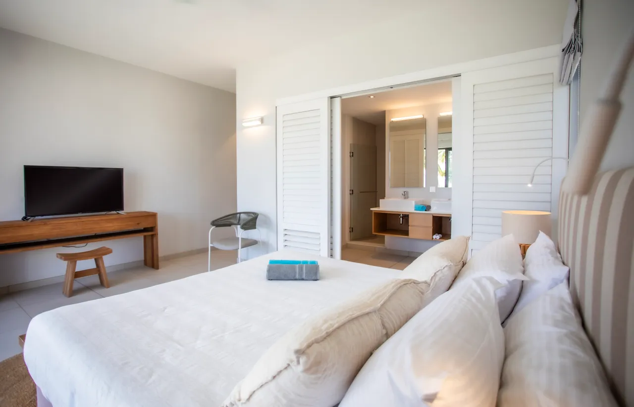 Chambre avec lit double et télévision pour un divertissement moderne.  

Éclairage indirect soulignant les teintes neutres des draps.  

Sol carrelé clair offrant une ambiance aérée et lumineuse.  

Mobilier simple en bois ajoutant une touche naturelle à l'espace.  

Accès direct à une salle de bain attenante pour plus de commodité.