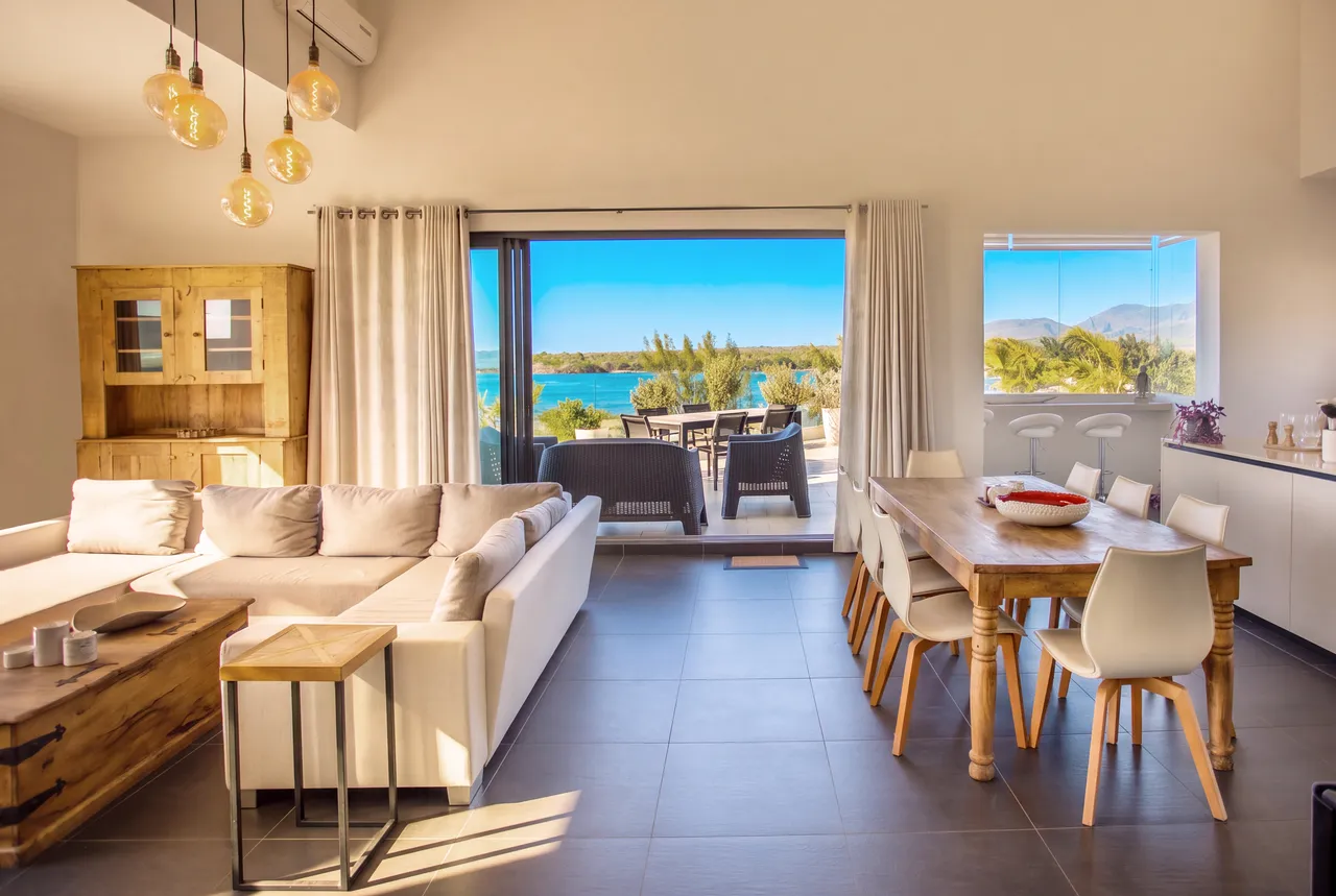 Tons neutres et naturels dominent dans un espace lumineux. Le salon dispose d'un canapé confortable et d'une vue sur la mer. Une grande table en bois invite à des repas conviviaux. Des fenêtres panoramiques apportent une lumière naturelle abondante.