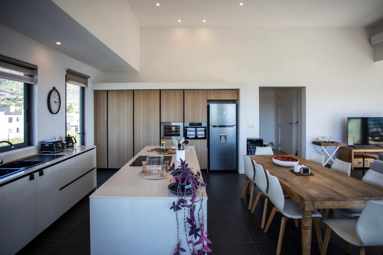 Couleurs claires et matériaux contemporains. Cuisine ouverte équipée avec îlot central et rangements intégrés. Salle à manger adjacente avec table en bois et chaises modernes. Lumière naturelle accentuée par de grandes fenêtres.