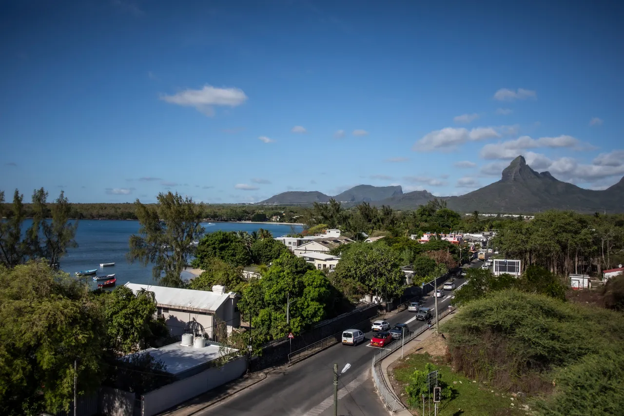 Vue panoramique sur les montagnes et la mer depuis la galerie. Route bordée d'arbres et habitations à proximité.