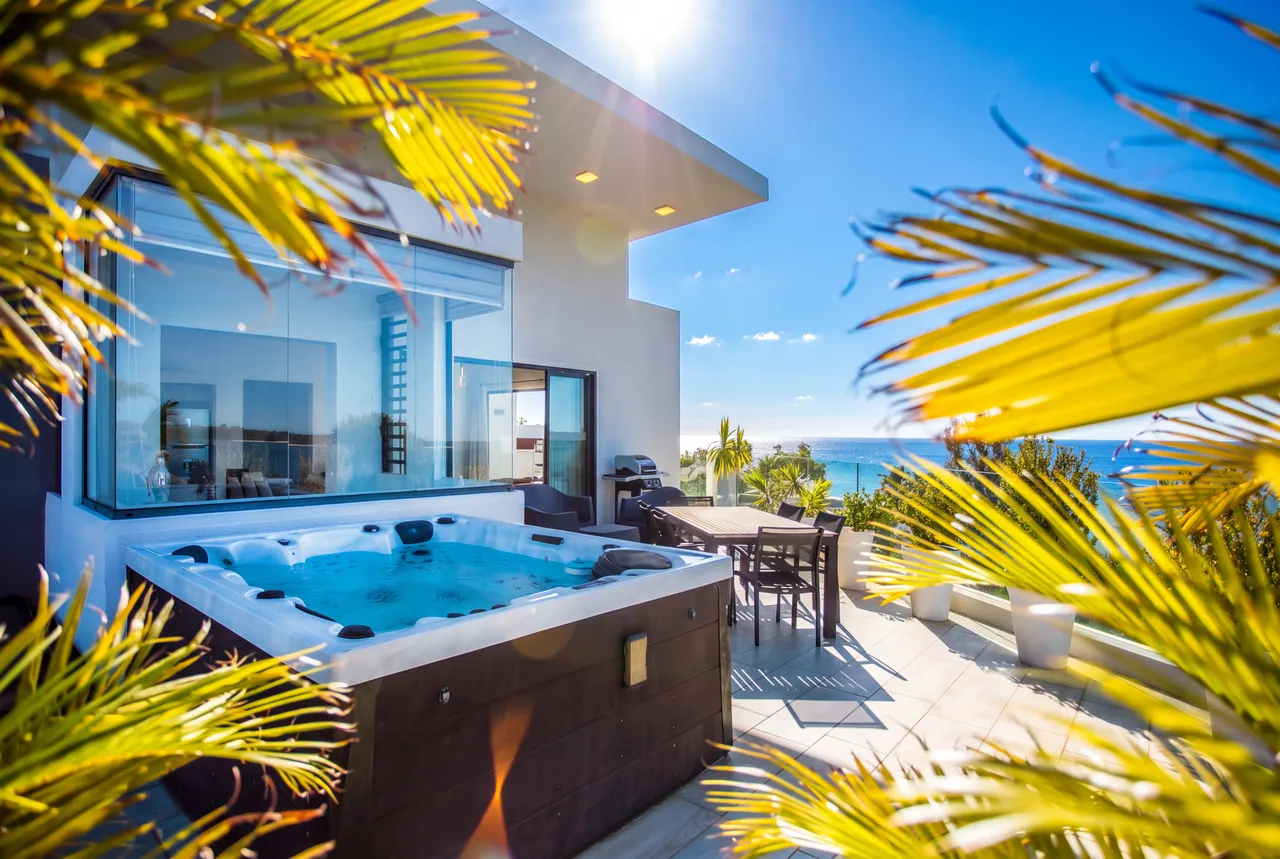 Terrasse ensoleillée avec jacuzzi, entourée de verdure et vue sur la mer. Table extérieure pour repas en plein air.