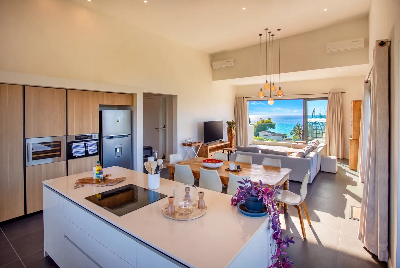 Sol en carrelage gris, cuisine moderne avec des finitions en bois clair. Vue sur la mer depuis le salon lumineux.