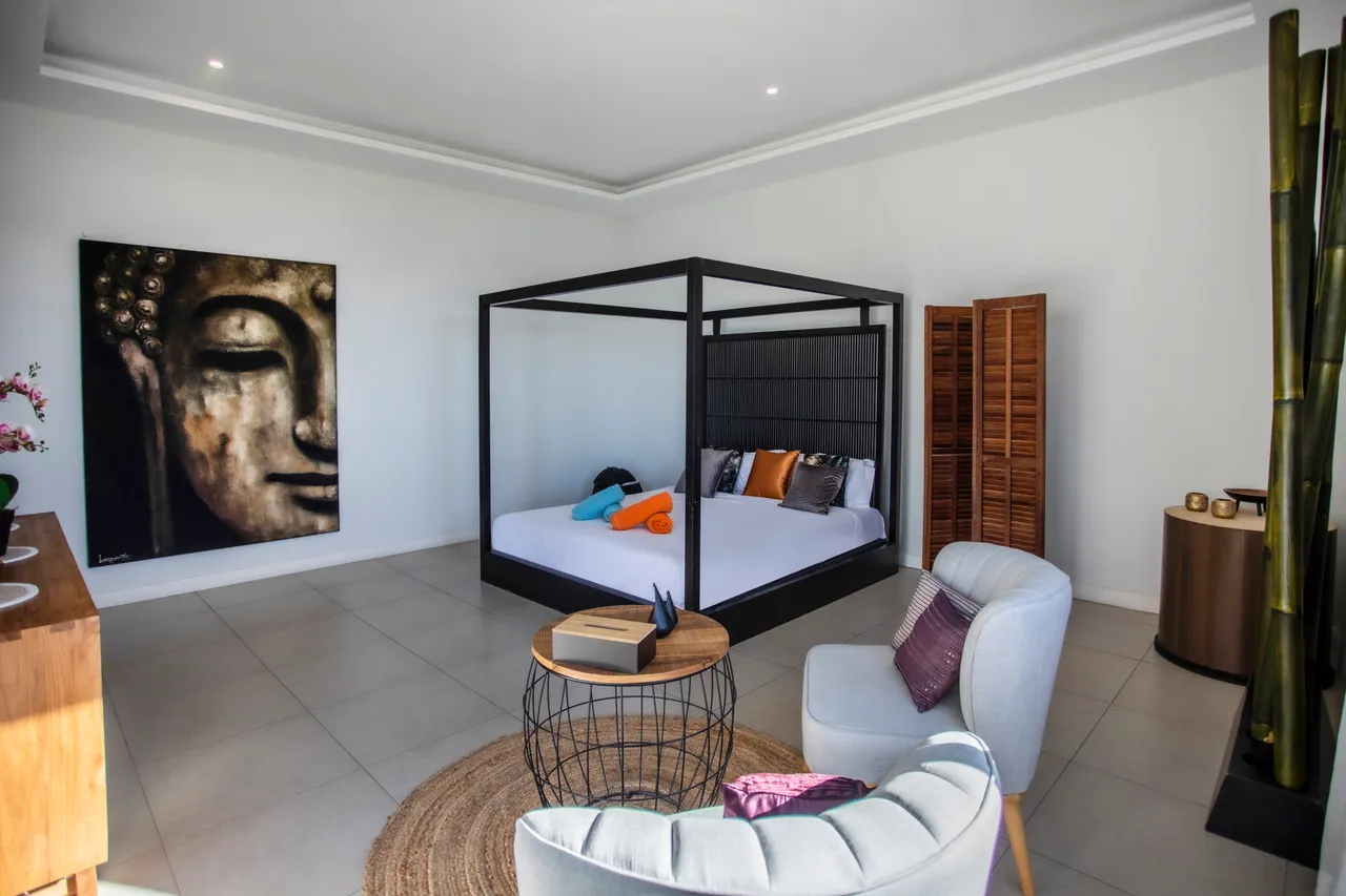 Couleurs neutres et éléments en bois ajoutent une touche chaleureuse. Chambre avec un lit à baldaquin, accompagnée d'un coin salon. Table en bois près d'une œuvre d'art murale. Rangement en bois intégré, décoré avec des tissus colorés. Éclairage naturel par de grandes fenêtres.