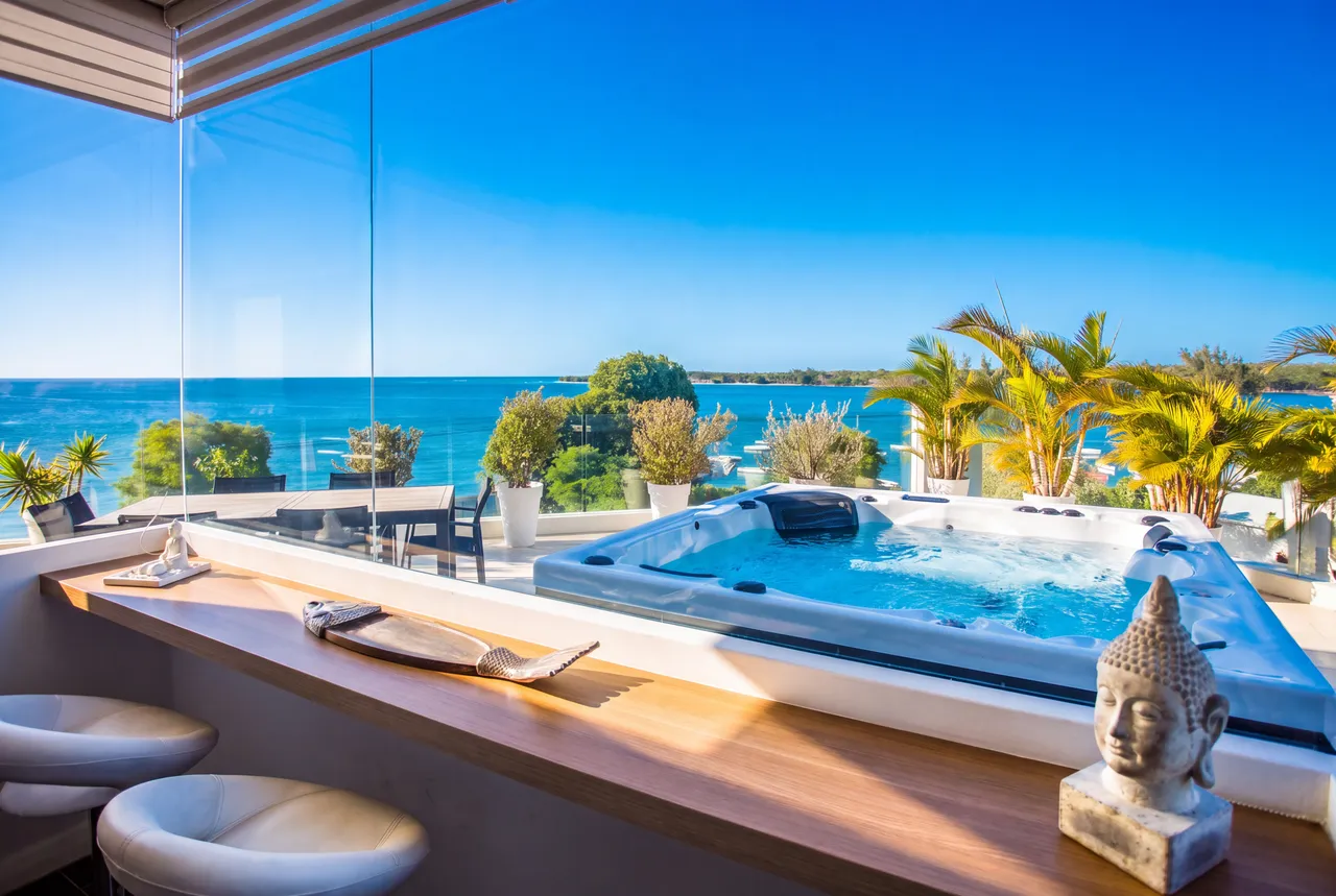 Bar en bois avec sièges modernes et vue sur la mer. Jacuzzi adjacent entouré de verdure.
