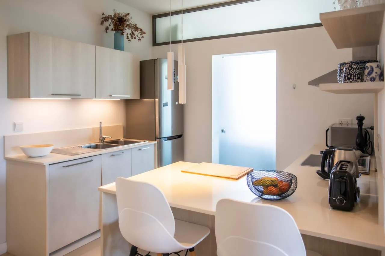 Teintes claires et surfaces lisses dominent la cuisine équipée. Bar à petit-déjeuner avec chaises modernes et éclairage suspendu. Électroménagers intégrés et plans de travail fonctionnels pour la préparation des repas. Aperçu de la salle d'eau depuis la cuisine.