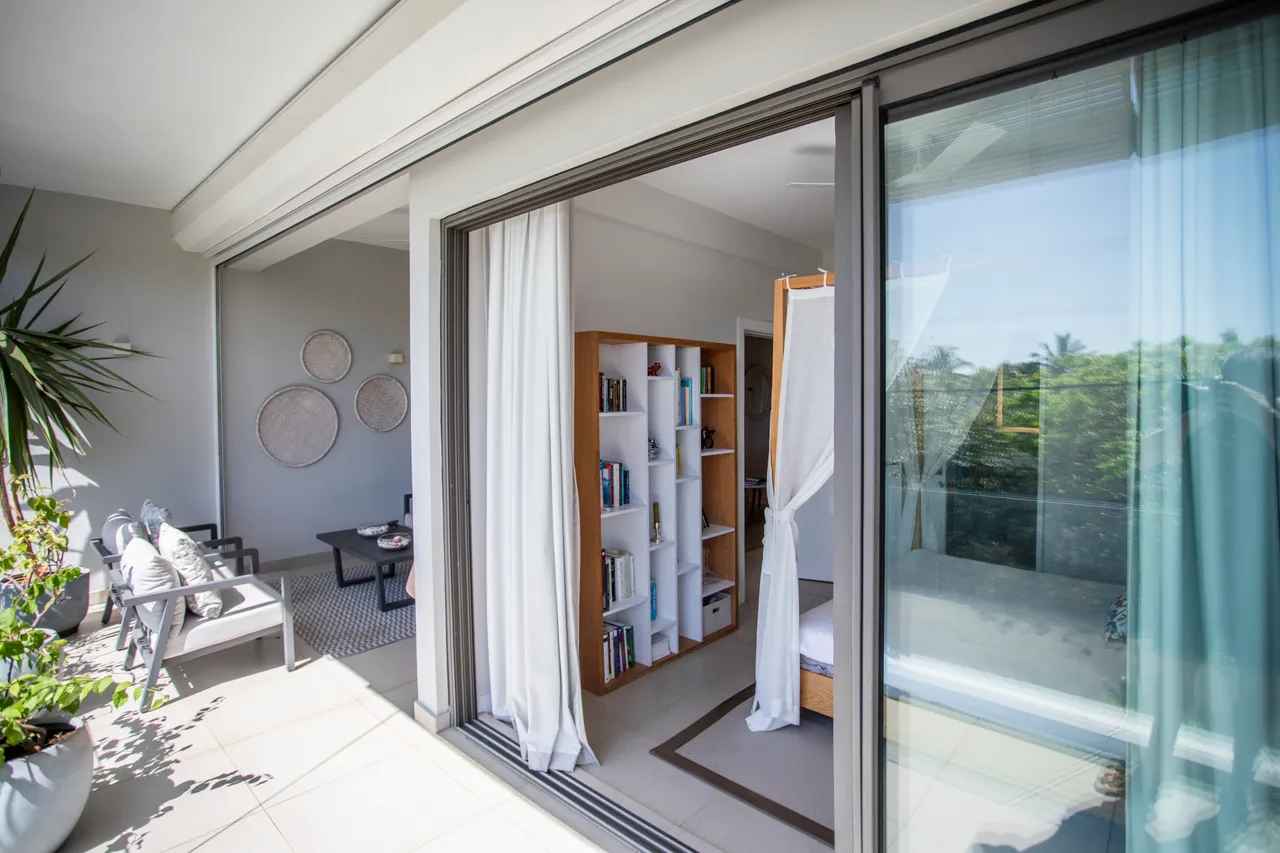 Balcon avec mobilier extérieur et vue sur la verdure environnante. Étagères avec livres ajoutent une touche personnelle. Sol en carrelage facile d'entretien. Rideaux légers laissant passer la lumière naturelle.