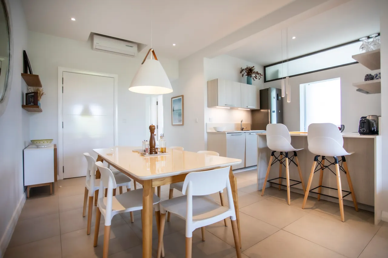 Lumière naturelle entrant par la fenêtre éclaire l'espace repas. Table en bois clair entourée de chaises blanches modernes. Cuisine ouverte avec bar et rangements pratiques. Éléments fonctionnels et agencement agréable.