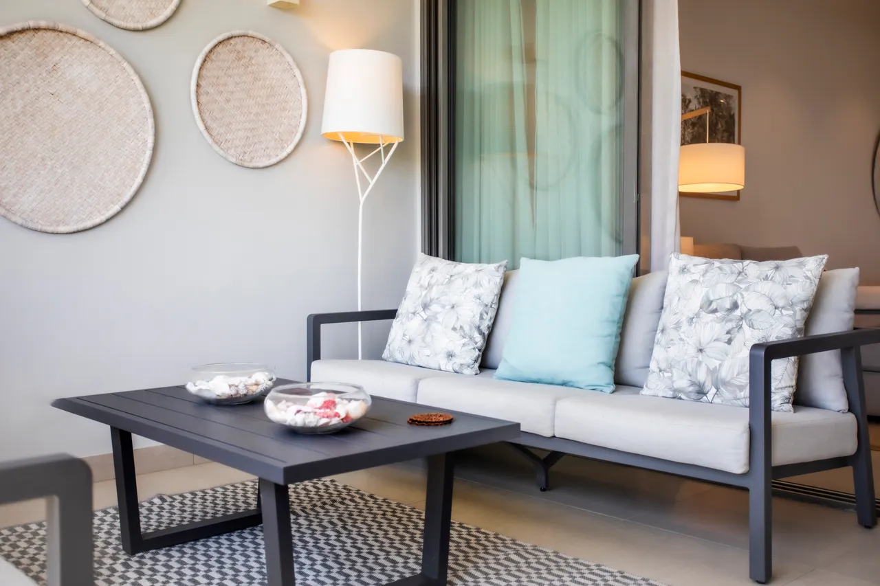 Lumière douce provenant d'une lampe sur pied, créant une ambiance apaisante. Canapé aux coussins fleuris accompagné d'une table basse en noir. Décoration épurée et éléments naturels sur les murs.