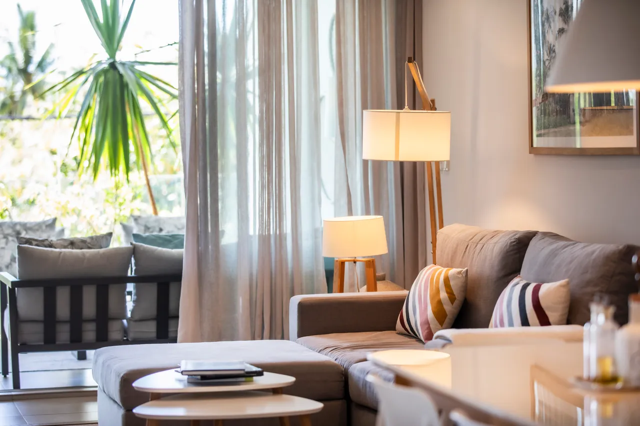 Salon lumineux avec grandes fenêtres, offrant une vue sur la végétation. Canapé et tables basses créent un espace convivial. Lampes fournissent un éclairage doux et chaleureux.