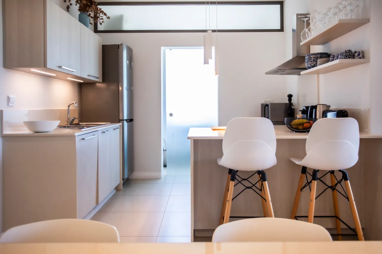 Cuisine moderne avec bar à petit-déjeuner et chaises blanches. Équipements intégrés et plans de travail clairs. Lumière naturelle grâce à une fenêtre sur l'extérieur. Rangements pratiques et agencement fonctionnel.