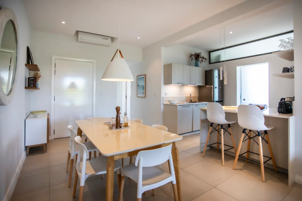 Espace repas avec table rectangulaire et chaises blanches modernes. Éclairage suspendu au-dessus de la table. Cuisine ouverte avec accès direct à l'extérieur.