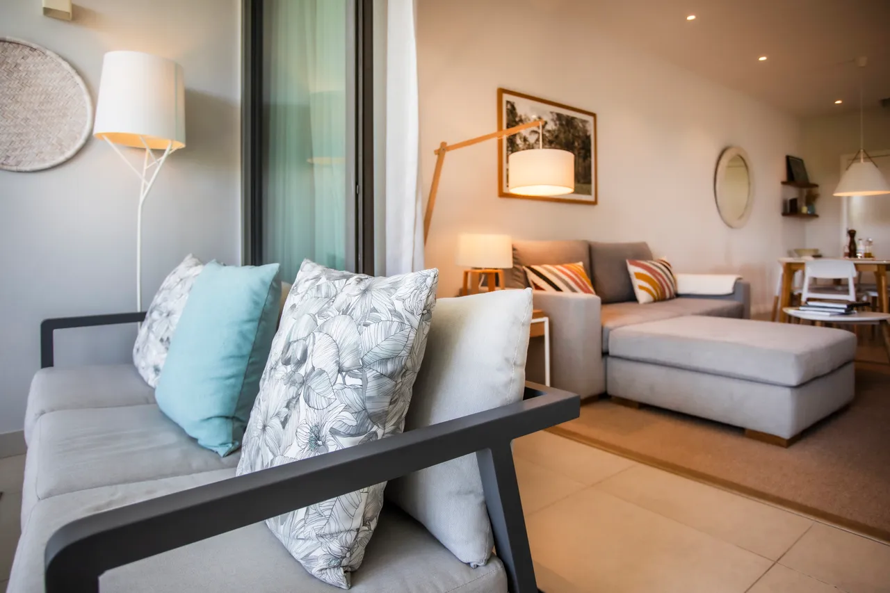 Canapé avec coussins décoratifs et éclairage doux dans le salon. Mobilier moderne et agencement ouvert créent une atmosphère accueillante. Sol en carrelage uniforme et clair.