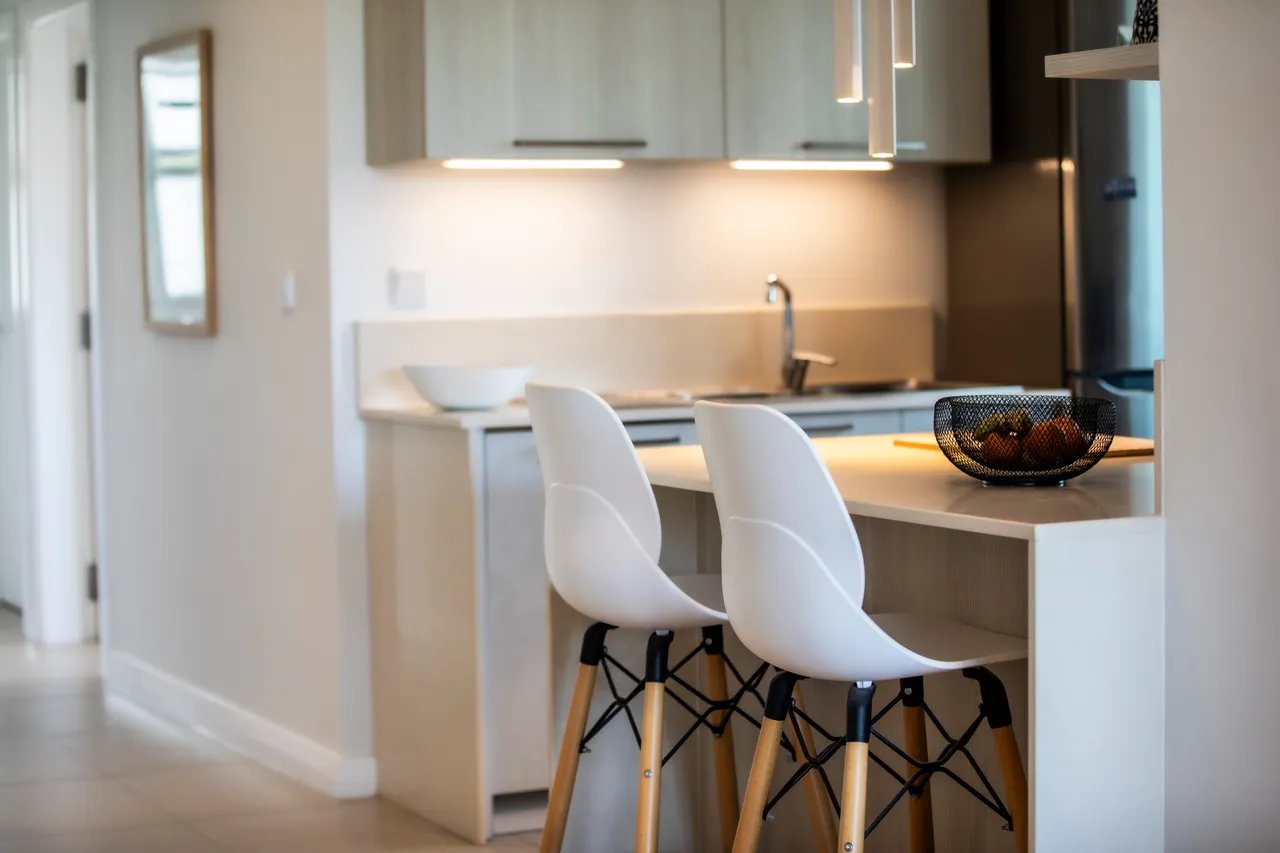 Cuisine contemporaine avec bar à petit-déjeuner et chaises blanches en bois. Éclairage intégré et électroménagers modernes.