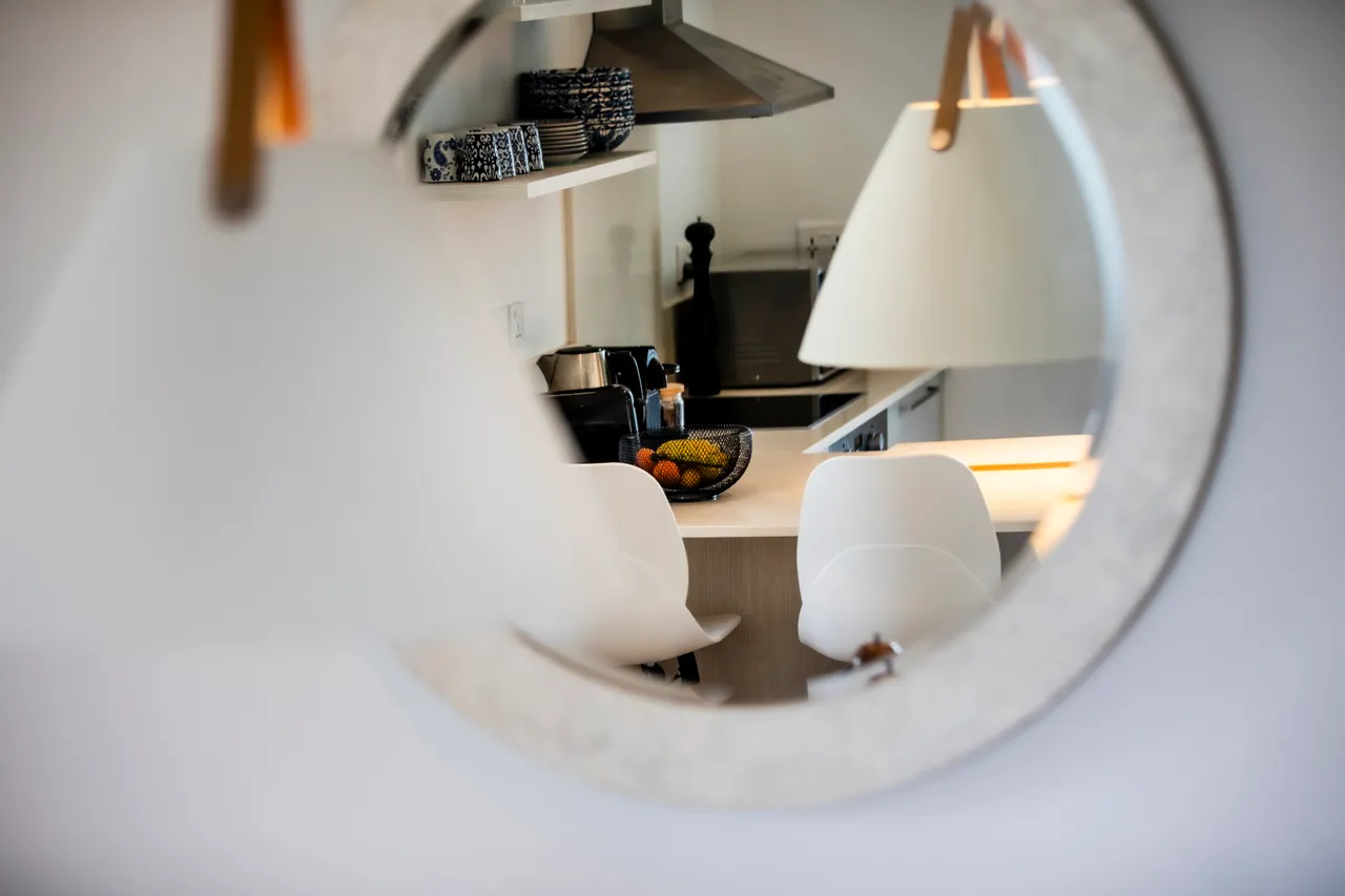 Miroir rond reflétant un coin cuisine avec bar et chaises modernes. Éclairage doux mettant en valeur l'espace. Équipements pratiques et décoration épurée.