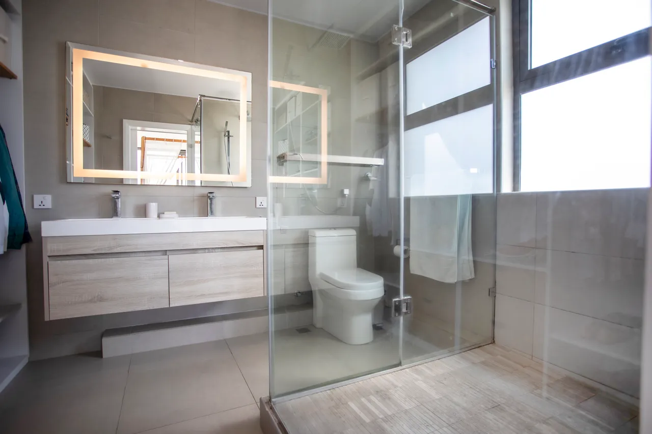 Salle de bain avec douche vitrée et meuble de rangement en bois. Éclairage moderne au-dessus du miroir.